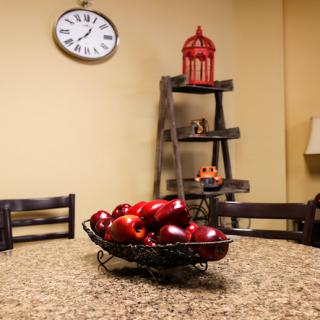 A cozy kitchen scene featuring a bowl of red apples on a granite table, complemented by a wall clock and decorative shelves.