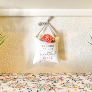 A small cushion with a 'Welcome to Our Comfortable Home' message hangs on the wall above a floral patterned surface.