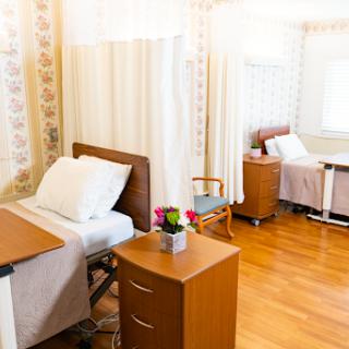 A well-lit hospital patient room featuring two beds separated by curtains, with a small table and flower arrangements.