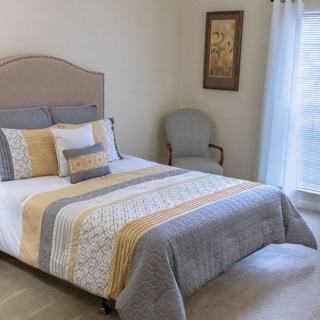 A neatly decorated bedroom with a bed, upholstered chair, artwork on the wall, and light coming through a window.