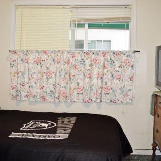 A cozy bedroom with a floral curtain and a bed covered in a black blanket, adjacent to a television on a dresser.