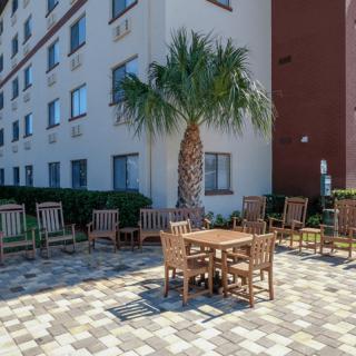 A patio area with wooden tables and chairs next to a building, featuring a palm tree and a paved surface.