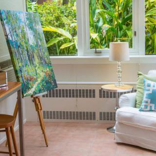 A bright art studio with a painting on an easel near a comfortable chair and lamp, overlooking lush greenery through large windows.