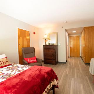 A cozy bedroom featuring a bed with a red blanket, an armchair, and wooden furniture on a wooden floor.