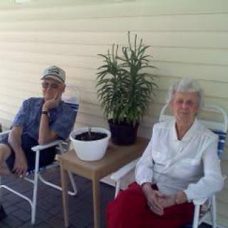 An elderly couple sitting on a porch, enjoying a relaxed moment together with potted plants nearby.