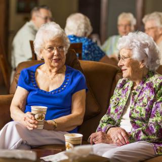 Two elderly women enjoy a conversation over coffee in a community setting.