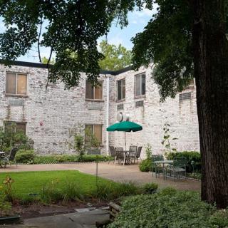 A serene courtyard with a stone building backdrop, shaded by trees and featuring outdoor seating with an umbrella.