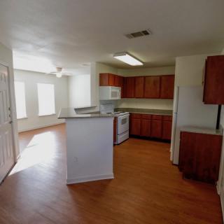 A bright kitchen with wooden cabinets and white appliances, featuring an open layout with ample natural light on a hardwood floor.
