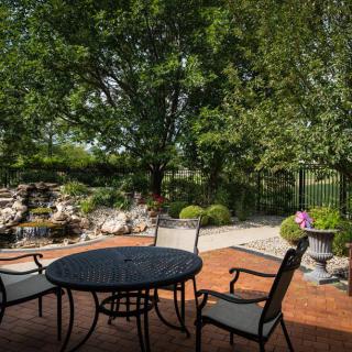 A serene patio setup with a round table and chairs, surrounded by lush greenery and a peaceful garden landscape.
