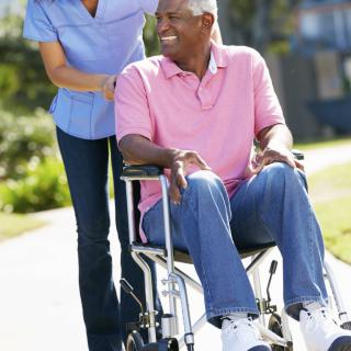 A caregiver assists an elderly man in a wheelchair outdoors.
