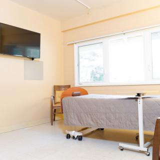 A clean and modern hospital room with a single bed, wall-mounted television, and large window.
