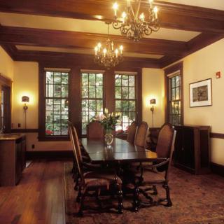 A traditional dining room with wooden furniture and chandeliers, featuring large windows and a floor rug.
