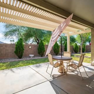 A sunny backyard patio with a table, chairs, and an umbrella set on a concrete floor surrounded by a lush garden.