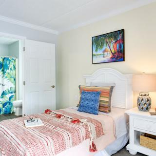 A cozy bedroom with tropical-themed decor, featuring a bed with colorful pillows and a patterned blanket, accompanied by a nightstand with a lamp and decorative items.