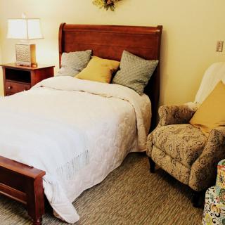 A cozy bedroom with a neatly made bed, pillows, an upholstered chair, and a lamp on a nightstand.