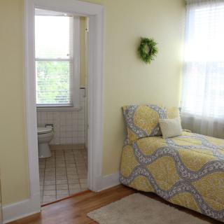 A cozy bedroom with a single bed featuring yellow bedding. The room has a doorway leading to a small bathroom, and large windows allowing natural light to fill the space.