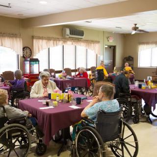Elderly residents gather in a nursing home dining area, seated around tables, enjoying a communal meal.