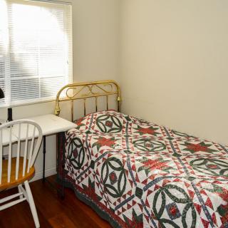 A cozy bedroom with a single bed adorned with a patterned quilt, next to a small desk and chair by the window.