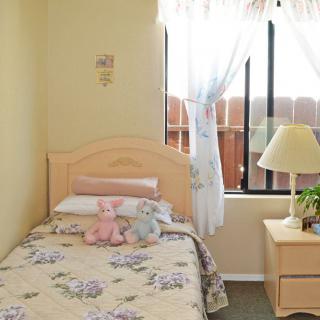 A cozy bedroom with a single bed, adorned with stuffed animals and floral bedding, next to a window letting in sunlight.