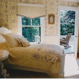 A cozy, traditionally decorated bedroom with a bed near a large window and an open door leading to an outdoor area.