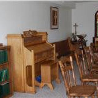 A small chapel with wooden chairs and an organ, featuring a cross and books on a shelf.