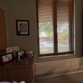 A cozy room with wooden blinds, showcasing a window view and a collection of photographs on a wooden dresser.