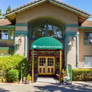 The entrance of a building with a green awning, surrounded by greenery on a sunny day.