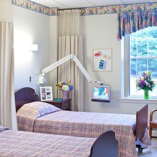 A hospital room with two patient beds, a window with flowers, and a mounted television.