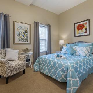 A cozy bedroom featuring a patterned bedspread, a comfortable chair, and artistic wall decor.