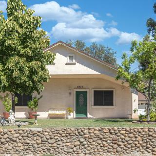 A quaint house with a green door, nestled behind a stone wall and surrounded by trees under a clear blue sky.