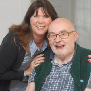 A woman is smiling as she poses with an elderly man indoors.