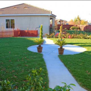 A well-maintained garden with a concrete pathway leads to a cozy house, featuring potted plants and lush green grass.