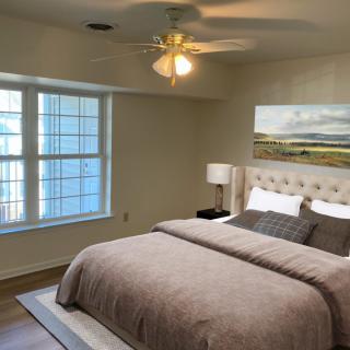 A cozy bedroom with a large window, featuring a neatly made bed and a ceiling fan.