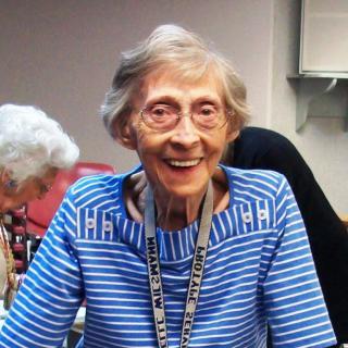 An elderly woman smiling brightly while wearing a blue and white striped shirt, sitting indoors with a name badge around her neck.