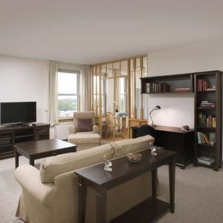 A cozy living room with a sofa, coffee table, and a television, featuring neutral colors and functional furniture such as a desk and bookshelf.