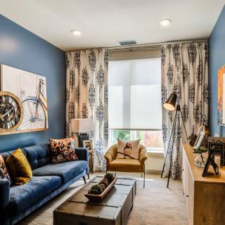 A modern living room with blue walls and stylish decor, featuring a sofa, artwork, and a large window with patterned curtains.