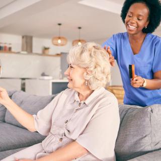 A caregiver assists a senior woman in her living room, both smiling and enjoying their time together.