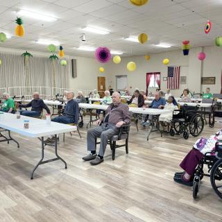 A community gathering of elderly people seated at tables in a decorated indoor space.