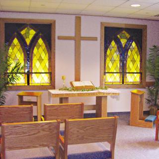 A small chapel featuring stained glass windows, an altar with a cross, and wooden chairs.
