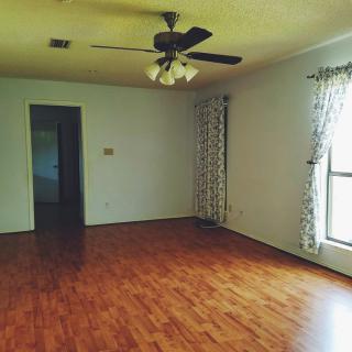 A spacious empty living room with wooden flooring, a ceiling fan, and large windows with curtains.