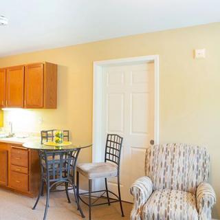 A cozy interior space featuring a small kitchenette with wooden cabinets, a high table with chairs, and a comfortable armchair.