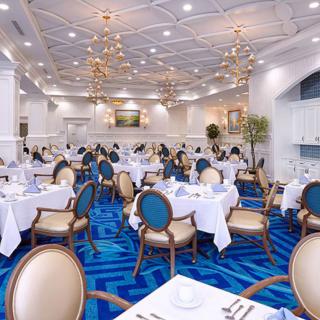 An elegant restaurant dining room with blue carpeting and white tablecloths, featuring classic seating and chandeliers.