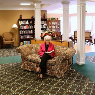 An elderly woman sits on a sofa reading in a cozy library setting.