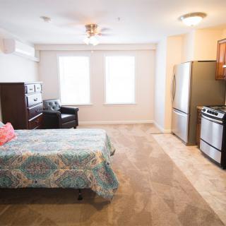 A well-lit studio apartment featuring a bed, kitchen area with stainless steel appliances, and ample natural light.