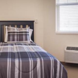 A neatly arranged bedroom with a single bed featuring striped bedding, next to a window and an air conditioner.
