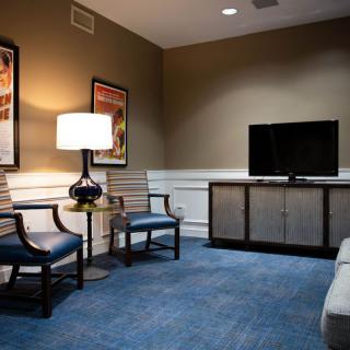 A cozy living room with a television, chairs, and a table lamp on blue carpet flooring.