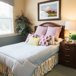 A cozy bedroom with a neatly made bed, decorated with multiple pillows, a nightstand with a lamp, and a framed picture on the wall.