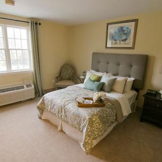 A cozy bedroom with a neatly made bed, decorative pillows, and a tray on top. The room features a window with curtains, an armchair in the corner, and artwork hanging above the headboard.