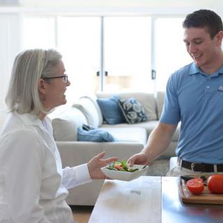 A caregiver serves a meal to a senior woman in a home setting.