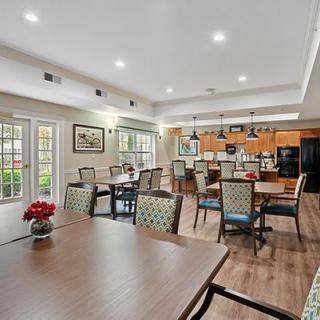 A spacious and well-lit dining area with a kitchen in the background, featuring multiple tables and chairs on a wood floor.